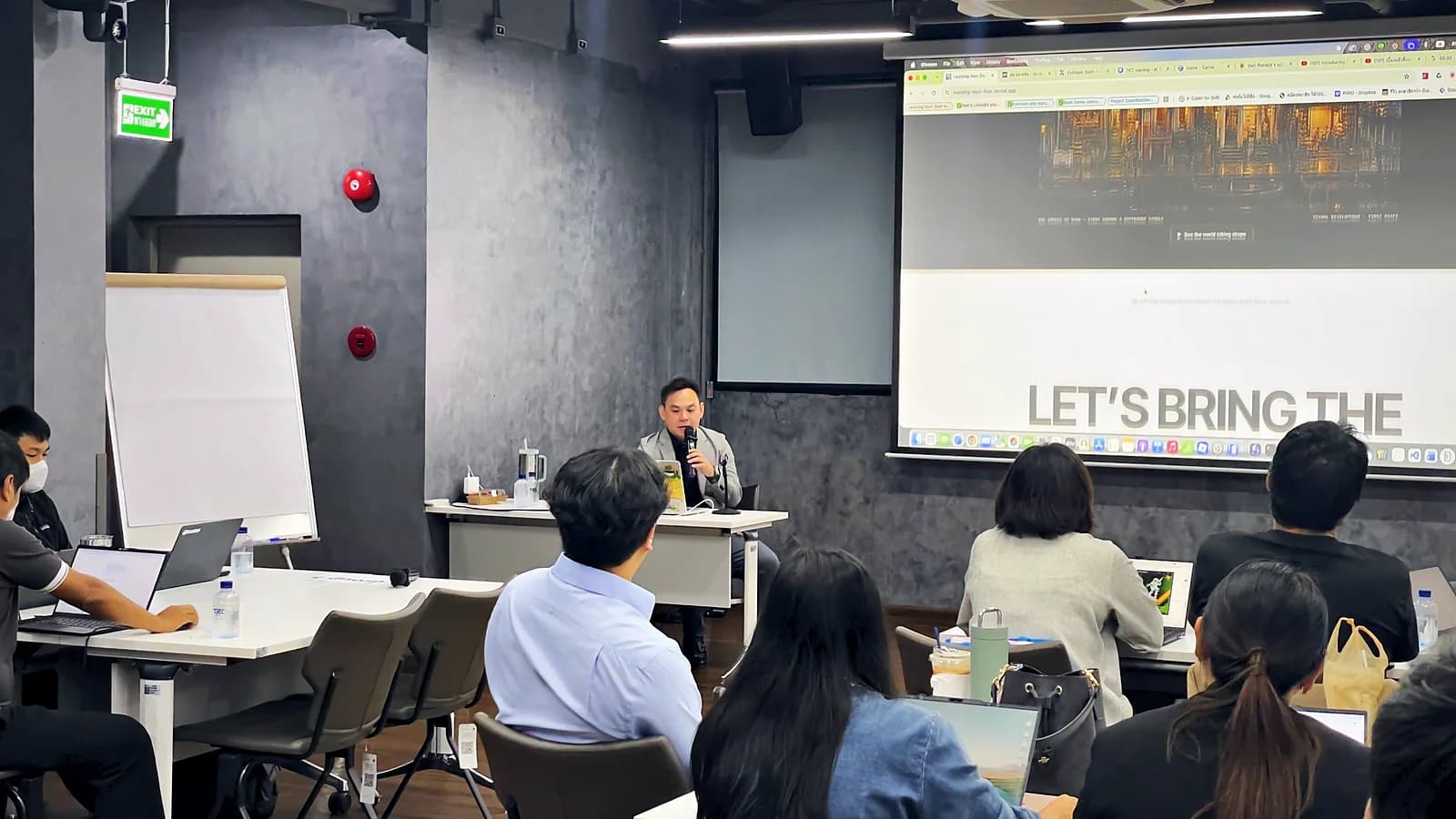 The speaker seated at the front with a microphone, presenting to participants with laptops